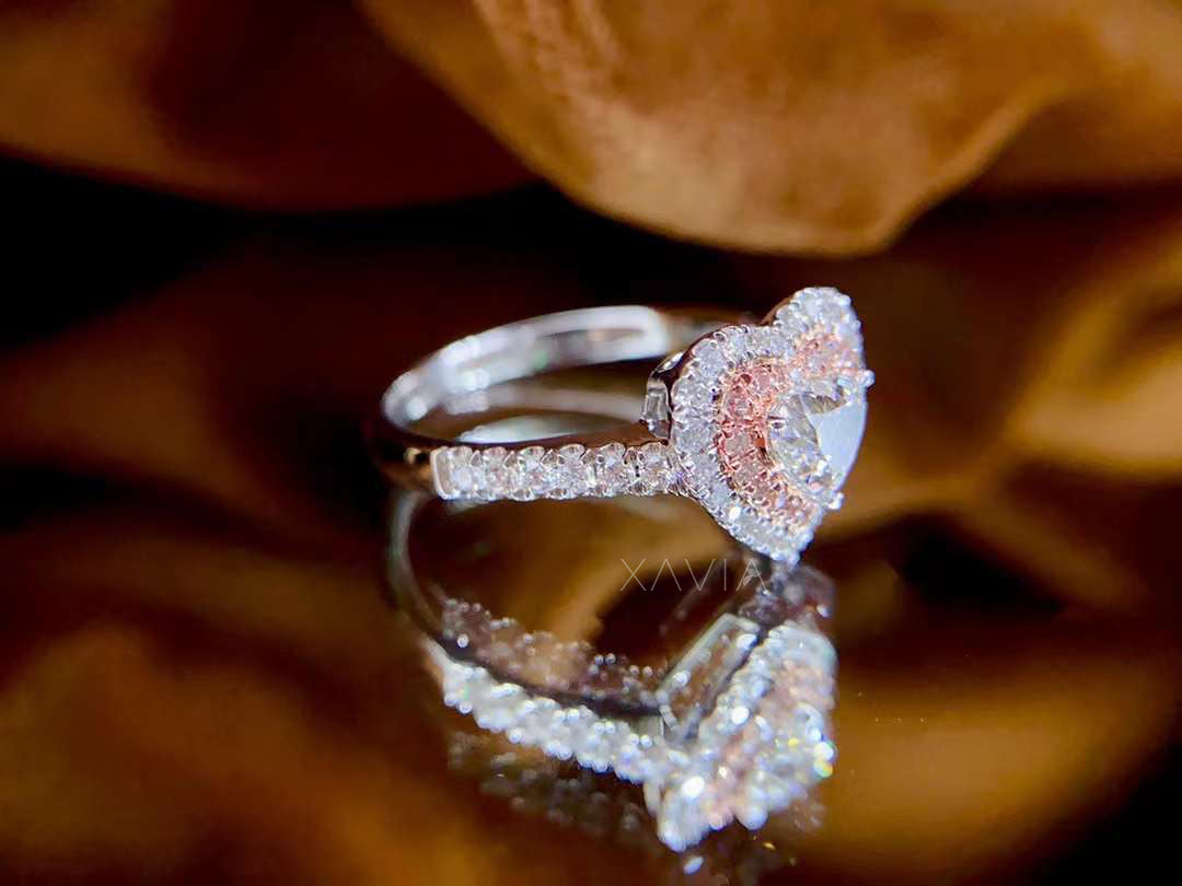 Angled side view of heart cut diamond ring with pink pavé halo and white diamond accents on pavé band, displayed on reflective surface