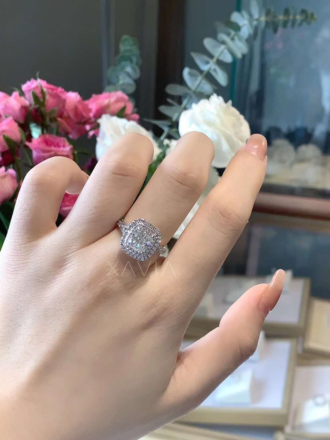 cushion cut double halo diamond engagement ring worn on hand with pavé band and floral backdrop