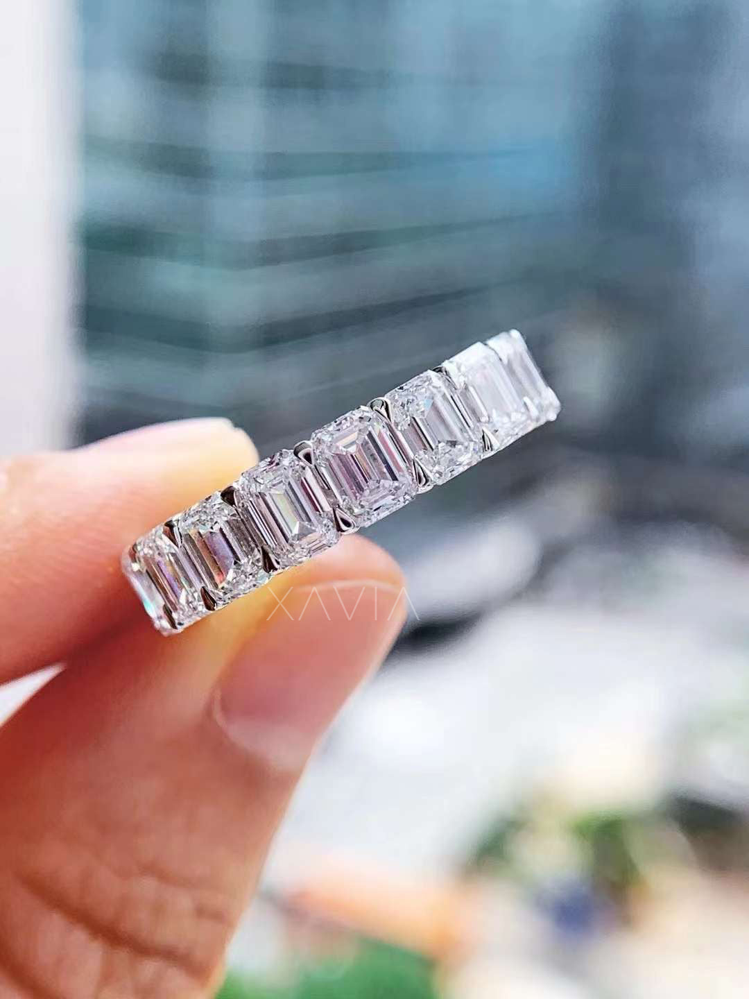 Macro close-up of the ring held delicately between fingers, emphasizing mirror-like flashes from the emerald cuts and prong detailing.