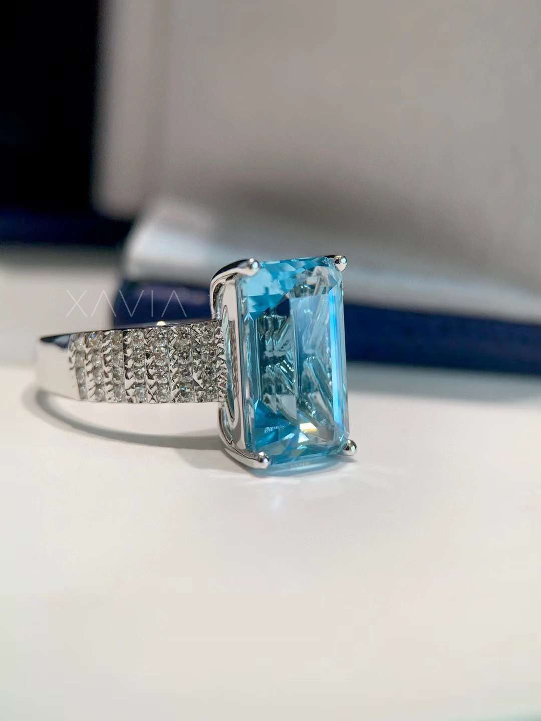 Side profile of emerald cut blue gemstone cocktail ring showing diamond pavé band and four-prong white gold setting in detail