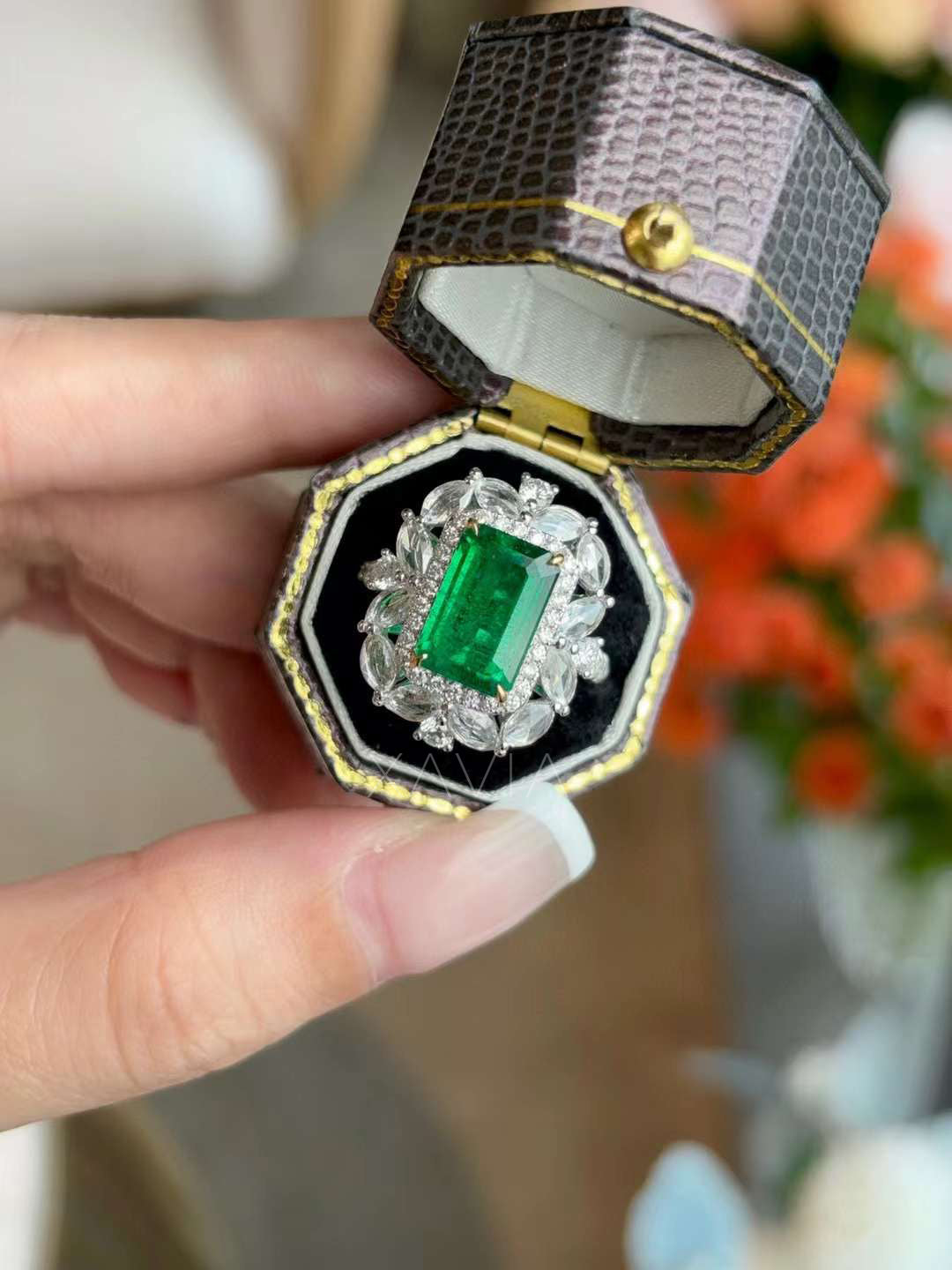 Luxury cocktail ring with emerald cut green gemstone center and marquise diamond floral halo photographed inside textured jewelry presentation box