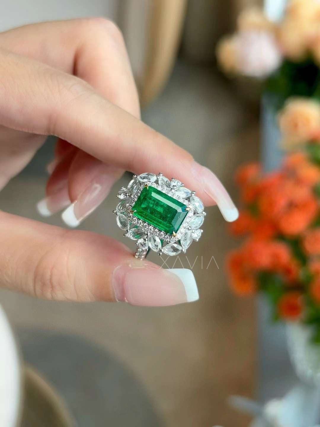 Handheld close view of emerald cut green gemstone ring surrounded by marquise and round diamond floral halo with pavé detailing in bright white metal