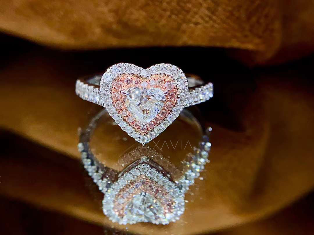 Luxury heart shaped diamond engagement ring with double halo design featuring pink and white pavé diamonds, displayed on reflective brown surface
