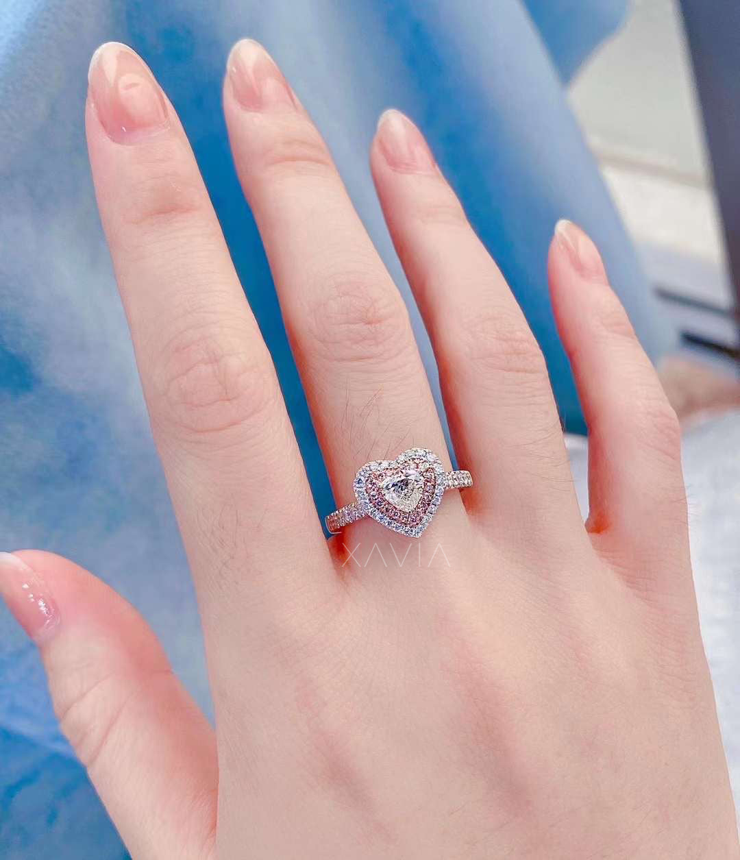 Heart cut lab-grown diamond engagement ring with pink pavé halo and outer white diamond halo, worn on hand against blue background