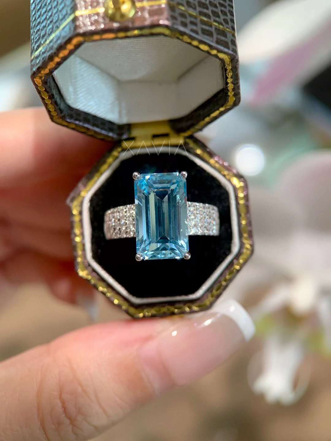 Luxury emerald cut blue gemstone ring with pavé diamond band displayed inside a black velvet jewelry box with gold trim