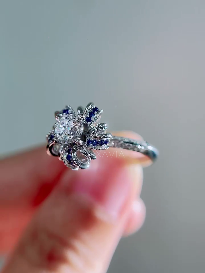 Hand held close perspective of round brilliant lab grown diamond ring with blue sapphire petal halo and pavé diamond band