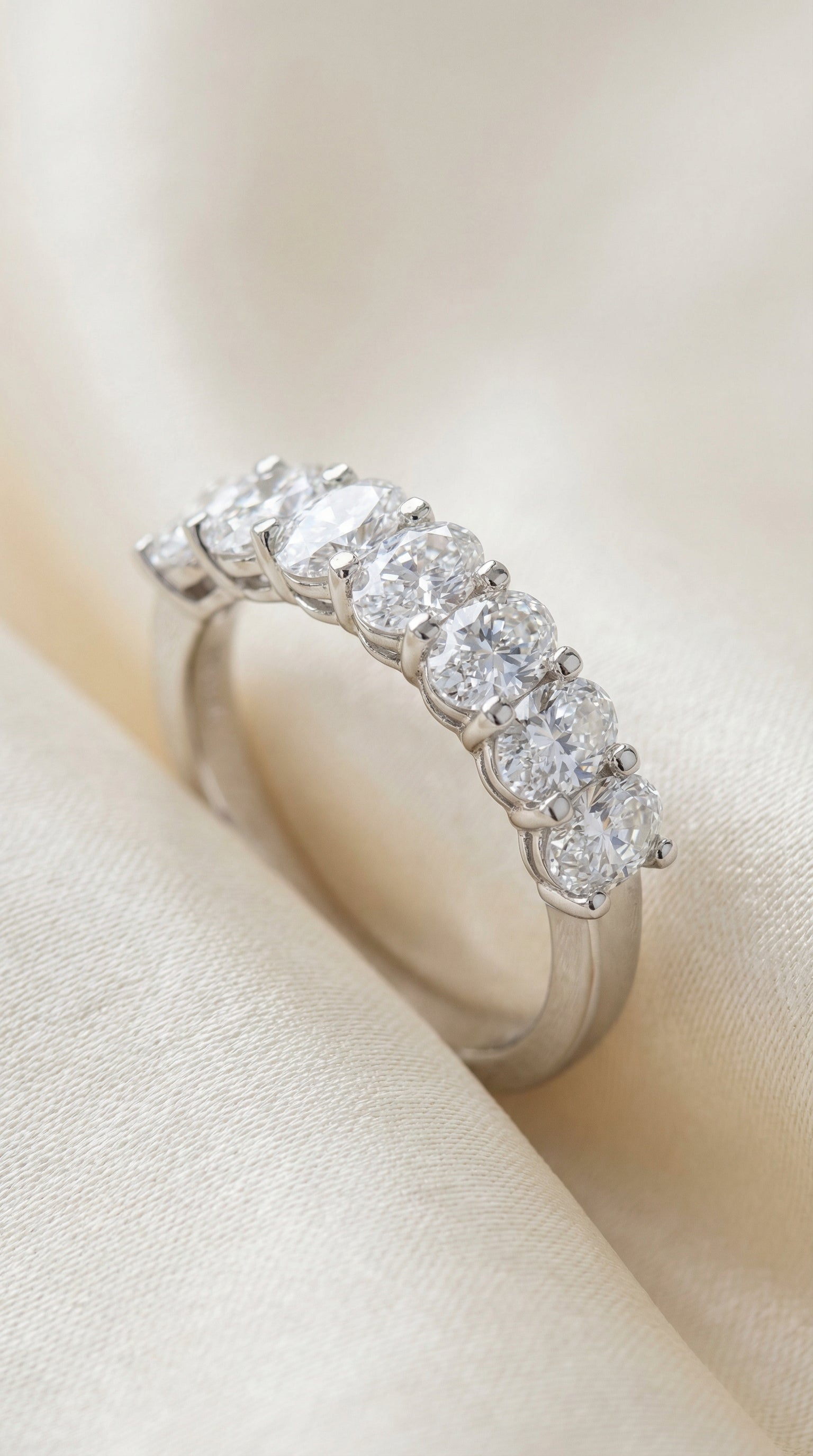 Close-up lifestyle image of oval lab grown diamond seven stone ring on textured fabric with flowers showing elegant prong set diamond row band