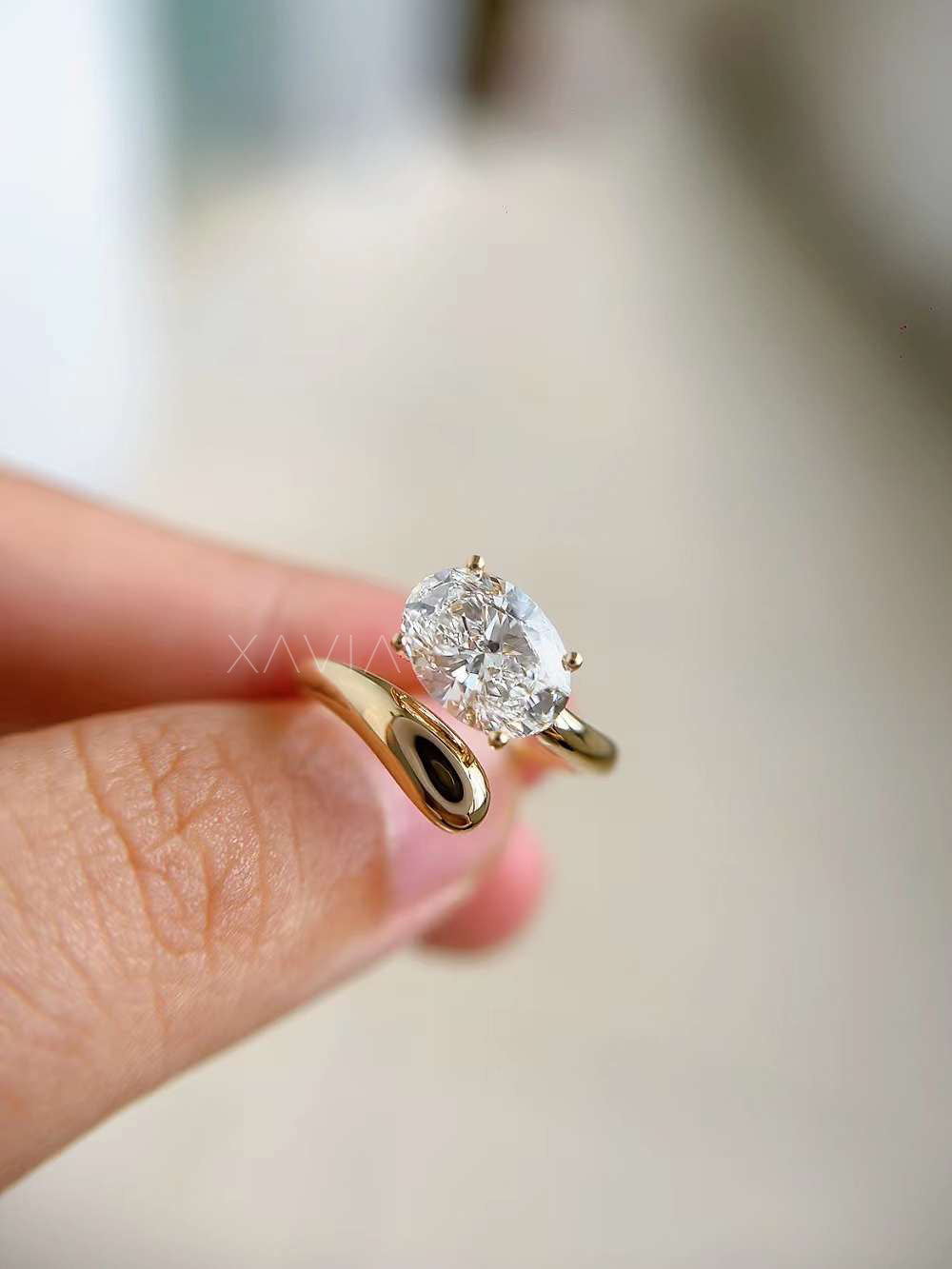 lifestyle shot of oval cut lab diamond open wrap bypass ring in polished yellow gold, worn on model’s hand with long manicured nails, modern minimalist engagement style over open book