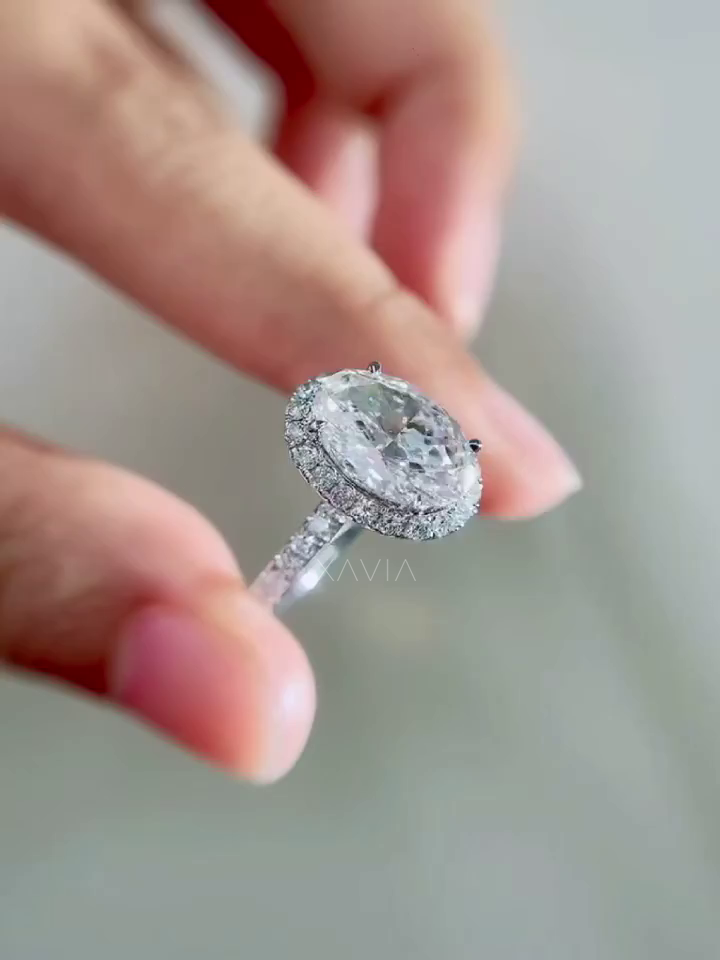 angled close view of oval cut lab grown diamond halo engagement ring with pavé halo and pavé diamond band held between fingers showing brilliant light reflection