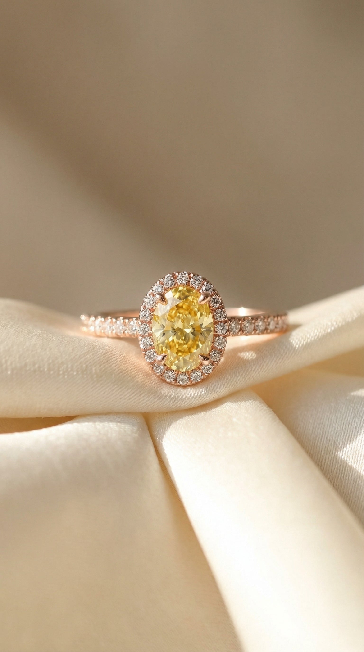 Oval yellow lab-grown diamond halo engagement ring with pavé band in rose gold displayed on silk fabric.