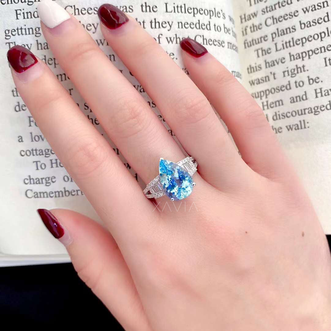 Pear cut aquamarine gemstone engagement ring worn on model hand showing large blue center stone with baguette diamond halo and pavé split shank design