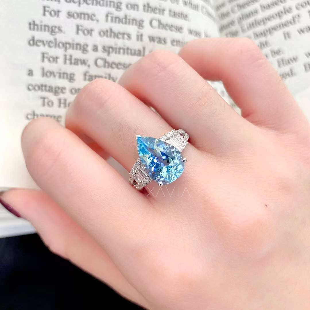 Angled view of pear shaped aquamarine diamond halo engagement ring on hand highlighting baguette side diamonds and pavé split shank brilliance