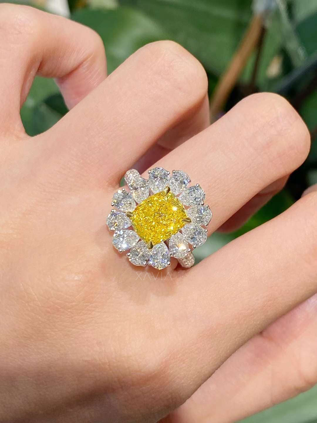 Ring with a yellow gemstone and diamond-like stones on a hand against a green leafy background