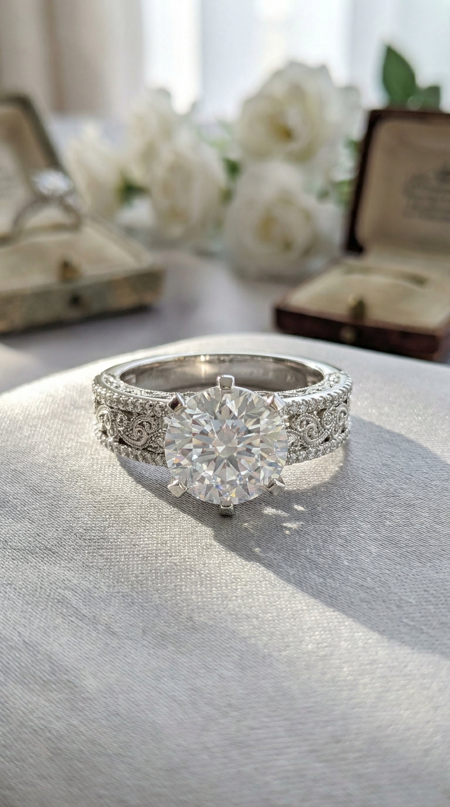 Vintage inspired round diamond engagement ring placed on satin fabric surrounded by pearls and dried flowers highlighting detailed filigree pavé band.